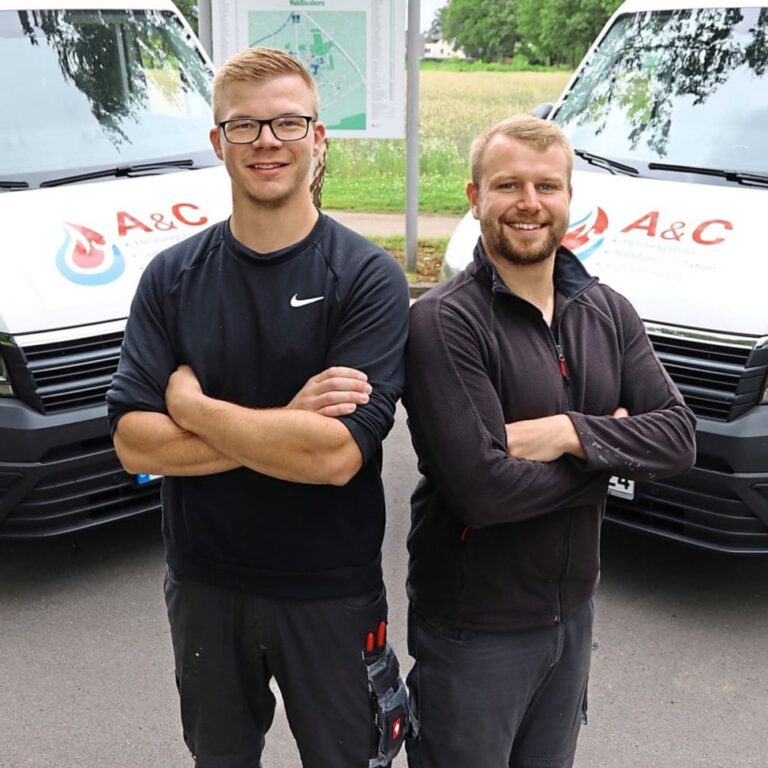 Zwei sympathisch lächelnde blonde Männer stehen mit verschränkten Armen vor zwei Transportern.