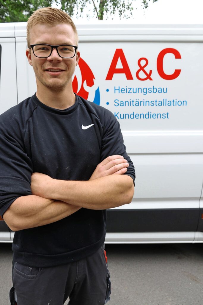 Ein sympathisch lächelnder blonder Mann mit einer Brille steht mit verschränkten Armen vor einem Transporter.